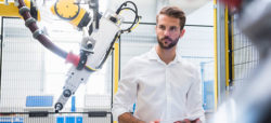 Na obrázku je muž v bílém košili, který se dívá na robota v průmyslovém prostředí. Robot má mechanickou ruku a je umístěn za ochrannou mříží. Muž drží tablet a působí soustředěně.