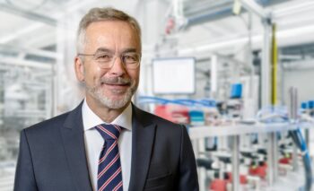 Muž ve formálním oblečení se usmívá na pozadí moderního výrobního prostředí.
