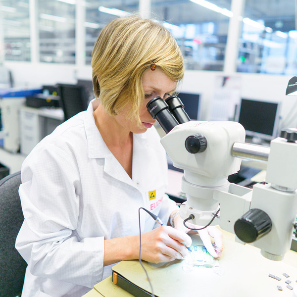 Žena v laboratoři pracuje pod mikroskopem, zkoumá malou součástku elektroniky. Nosí bílý plášť a jedná se o technologické prostředí s počítači v pozadí.