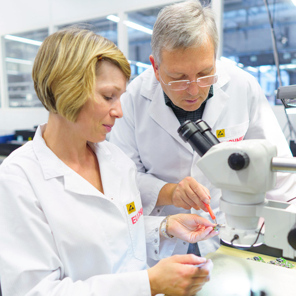 Na obrázku jsou dva pracovníci v laboratoři. Žena s krátkými vlasy a bílým pláštěm se dívá na malý objekt, který drží v ruce, zatímco muž s brýlemi se k ní sklání a ukazuje na něj. Oba работают в technickém prostředí s mikroskopem a dalšími vybaveními.