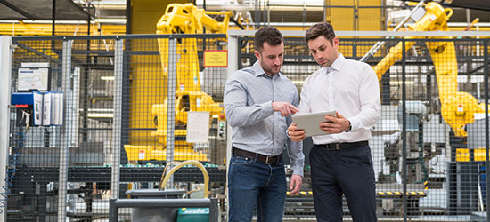 Zwei Männer stehen in einer modernen Industrieumgebung und betrachten gemeinsam ein Tablet. Im Hintergrund sind gelbe Industrieroboter und Maschinen sichtbar.