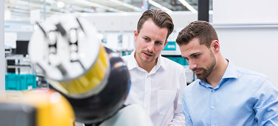 Zwei Männer in einem modernen Arbeitsumfeld betrachten aufmerksam eine Maschine oder technische Vorrichtung. Der Mann links trägt ein weißes Hemd, der rechts ein hellblaues. Im Vordergrund ist ein Teil der Maschine zu sehen.