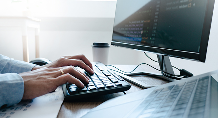 Eine Person tippt auf einer Tastatur, während ein Monitor mit Codezeilen im Hintergrund zu sehen ist. Neben dem Computer steht ein Kaffeebecher. Der Arbeitsplatz ist hell und modern eingerichtet.