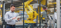 Ein Mann steht vor einer großen, metallenen Umzäunung in einer industriellen Umgebung und betrachtet ein Tablet in seinen Händen. Im Hintergrund ist ein gelber Roboterarm und weitere Maschinen sichtbar.