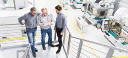 Three men in gray shirts stand on a staircase in a modern industrial setting, reviewing documents together. In the background, various machines and equipment are visible.