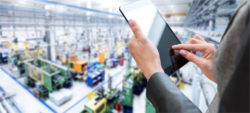 A person holds a tablet while in a spacious industrial environment with various machines and equipment in the background. The setting appears to be a factory or manufacturing site.